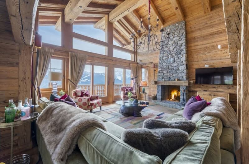 Spacious living room with high wooden ceilings, large windows offering a scenic mountain view, a stone fireplace, plush sofas and armchairs, and a chandelier lighting the cozy interior