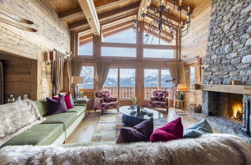 Spacious living room with high wooden ceiling, large windows offering a scenic mountain view, stone fireplace, plush sofas and armchairs with colorful patterned upholstery, and a chandelier lighting the cozy space.