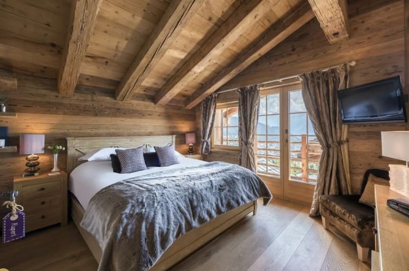 Rustic wooden bedroom with exposed beams, large windows overlooking a balcony or terrace, a king-size bed with a cozy gray comforter, and wooden furniture including nightstands and a TV stand.
