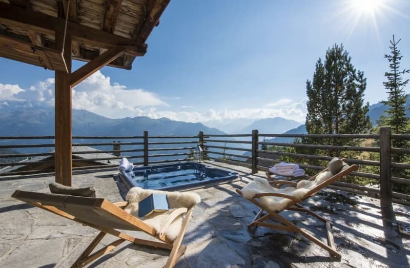 Wooden deck with a hot tub overlooking a scenic mountain landscape with pine trees and a blue sky with clouds. The deck has a wooden railing and is covered by a wooden roof structure.