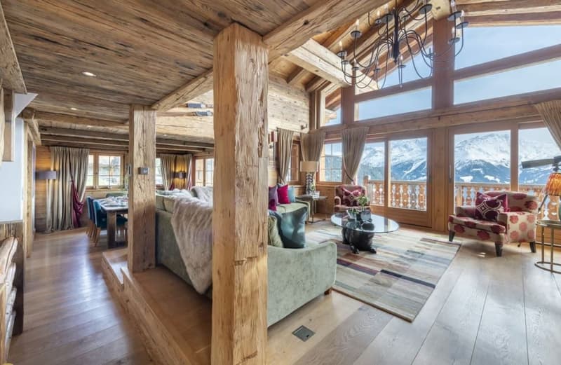 Spacious open-plan living area with wooden beamed ceiling, large windows offering mountain views, plush sofas and armchairs, a fireplace, and a chandelier lighting fixture.