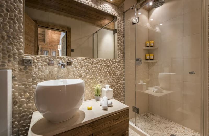 Rustic bathroom with stone wall tiles, wooden vanity, vessel sink, glass shower enclosure, and shelves with toiletries.