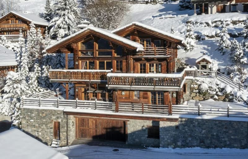 Two-story wooden chalet-style house with large balconies, surrounded by snow-covered trees and a garage.