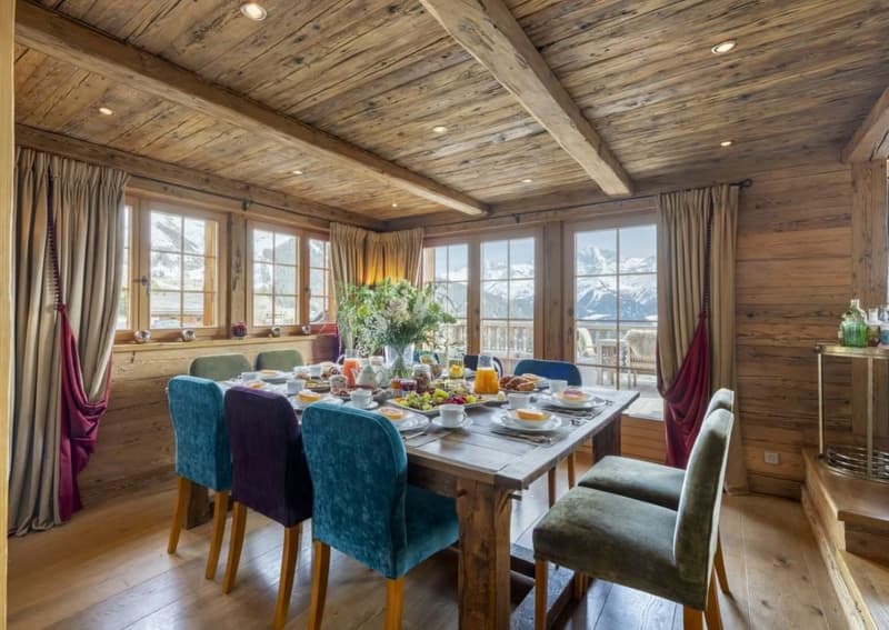 Rustic wooden dining room with large windows overlooking a scenic mountain view, wooden beamed ceiling, wooden dining table with colorful upholstered chairs, and a floral centerpiece on the table