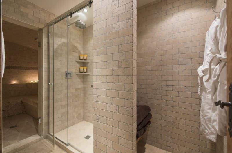 Large walk-in shower with glass enclosure, stone tile walls, shelves for toiletries, and a towel rack.