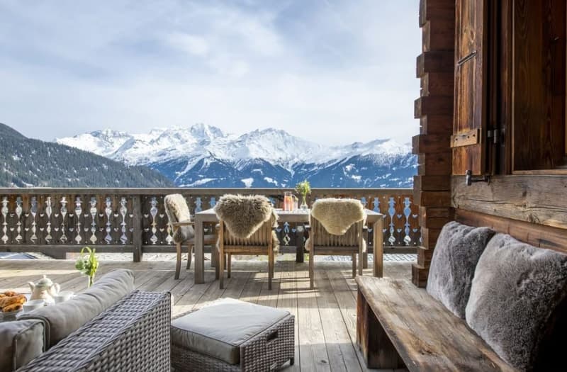 Wooden terrace with cozy seating area, including wicker chairs with fur throws, overlooking a scenic mountain landscape with snow-capped peaks in the distance.