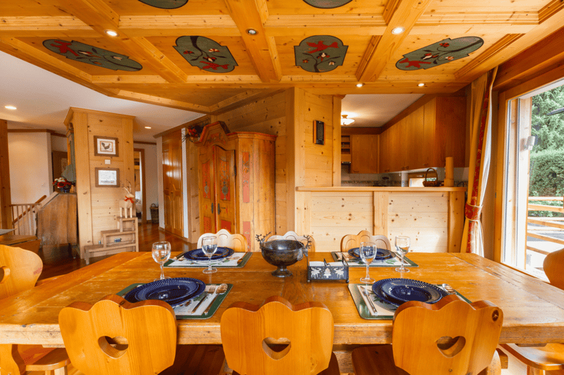 Large wooden dining table, four blue plates, six wine glasses, and wooden chairs with cut-out hearts. Wooden interior with large wooden cabinets, wooden flooring, and wooden ceiling beams. Open sliding glass doors to the kitchen. View of the outdoors.