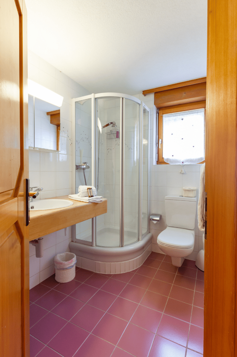Bathroom with shower cubicle, sink, mirror, toilet, and tiled floor. A white towel on the sink, a trash can, and a toilet paper holder. A window with white curtains.