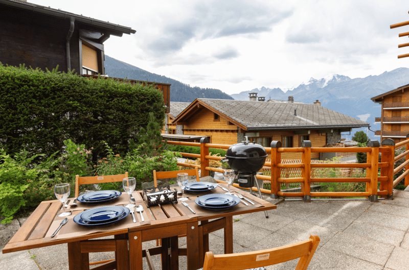 Wooden dining table set for four with plates, glasses, and cutlery, next to a grill, surrounded by plants and a fence, with a view of a mountain