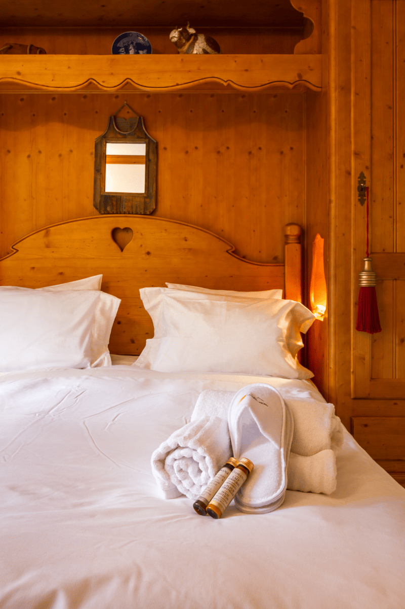 Large wooden bed with headboard, white bed sheets, white pillowcases, 2 bottles of lotion, 2 white towels, slippers, wooden shelf with mirror, wooden wall cabinets, wooden flooring