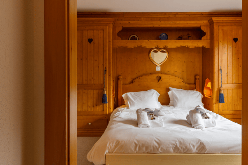 Bedroom with wooden walls and bed, wooden headboard, pillows, folded towels, and a heart-shaped shelf with items on top. Wooden cabinet with a door on the left side and a blue tassel hanging from a rod. On the right, there is a cabinet with a heart design