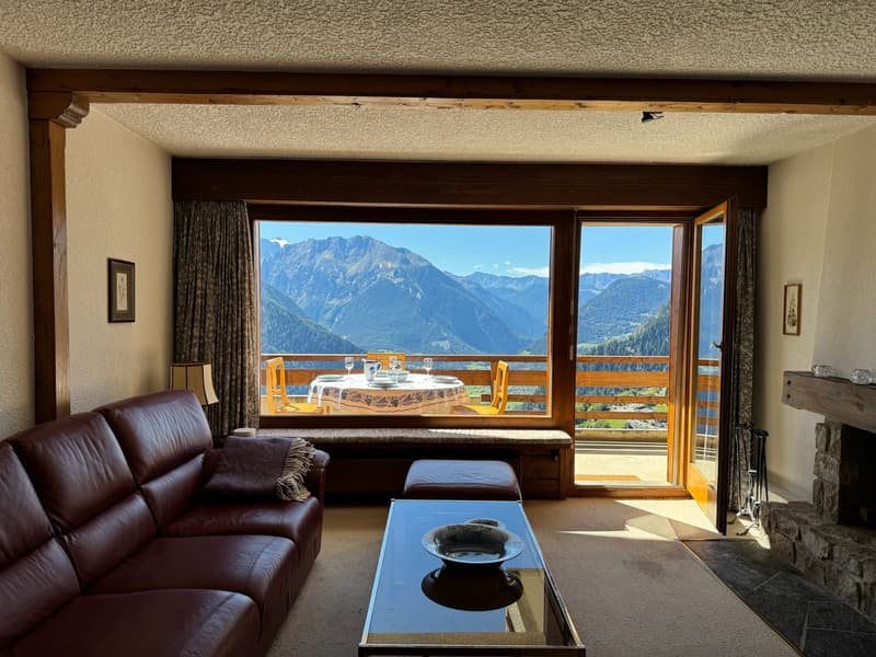 A large window overlooking the mountain range, a cozy fireplace, a coffee table with a bowl, and a leather couch.
