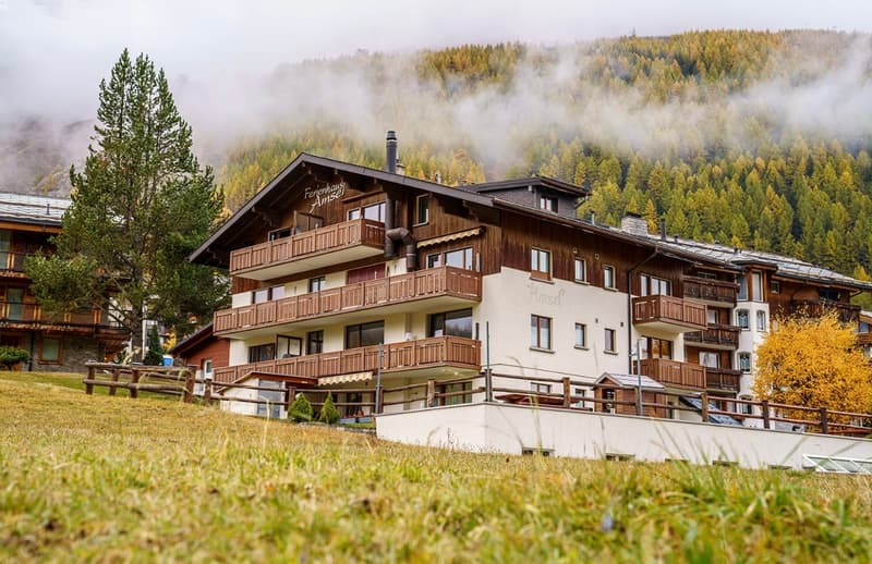 3-story building, brown balconies, brick and white wall, lush grass, pine trees, fog in the background, mountains