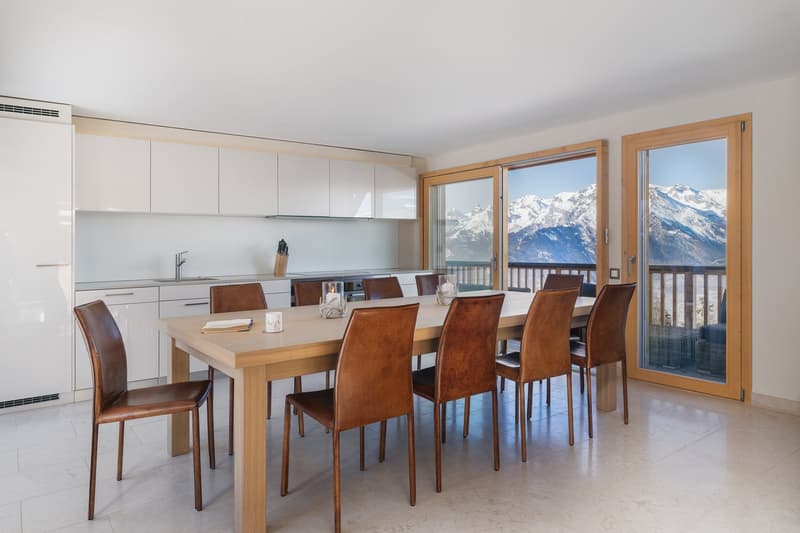 Dining room with a large wooden table, leather chairs, sliding glass doors with a balcony, kitchen, and a view of snowy mountains