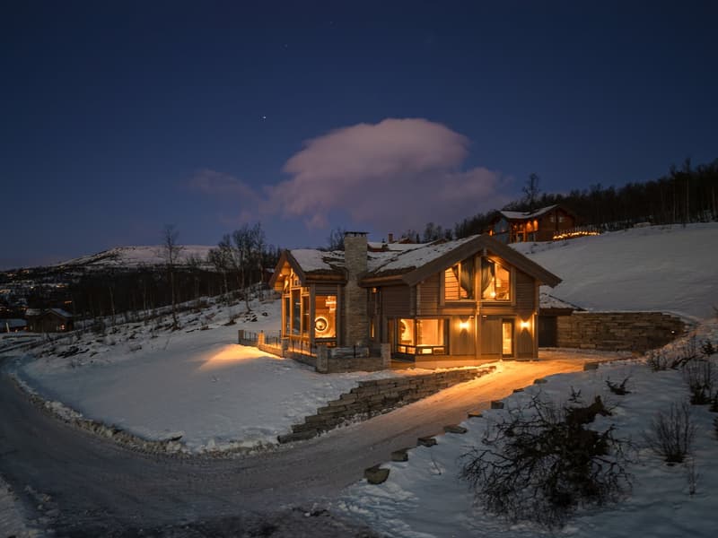 wooden house, covered in snow, stone foundation, glass windows, lit up at night, located on hillside