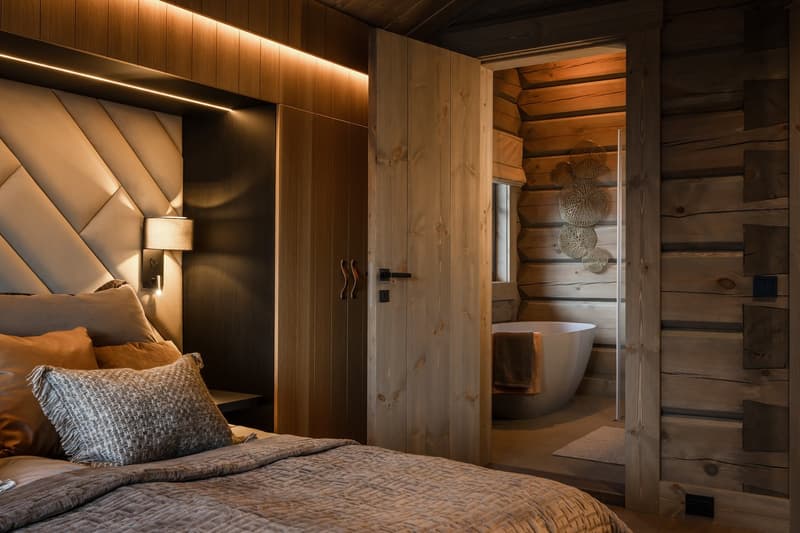 wooden walls, a bed with white, beige, and brown pillows, a white headboard with lights, a white bathtub with towels in the bathroom, a lamp