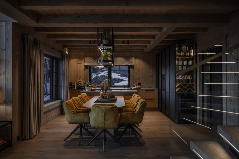 Dining table with chairs, wooden wall, plants, lamp, kitchen area, wine rack, view of snow-covered mountains through the windows