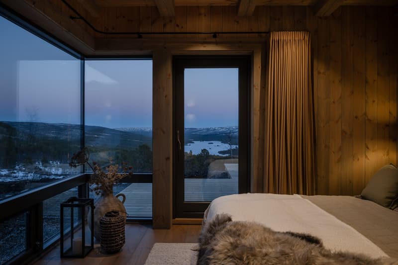 A bedroom with a bed, wooden walls, large windows, and a balcony with a view of a lake and mountains