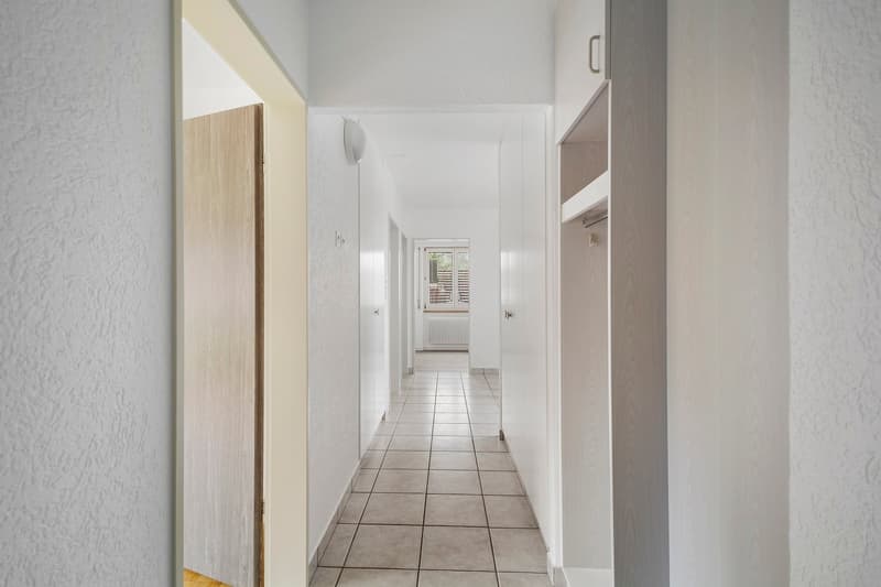 The image shows a long, narrow hallway with white walls and a tiled floor. The hallway has several doors leading to other rooms, and a window at the end providing natural light. The hallway appears to be part of a larger residential or commercial property