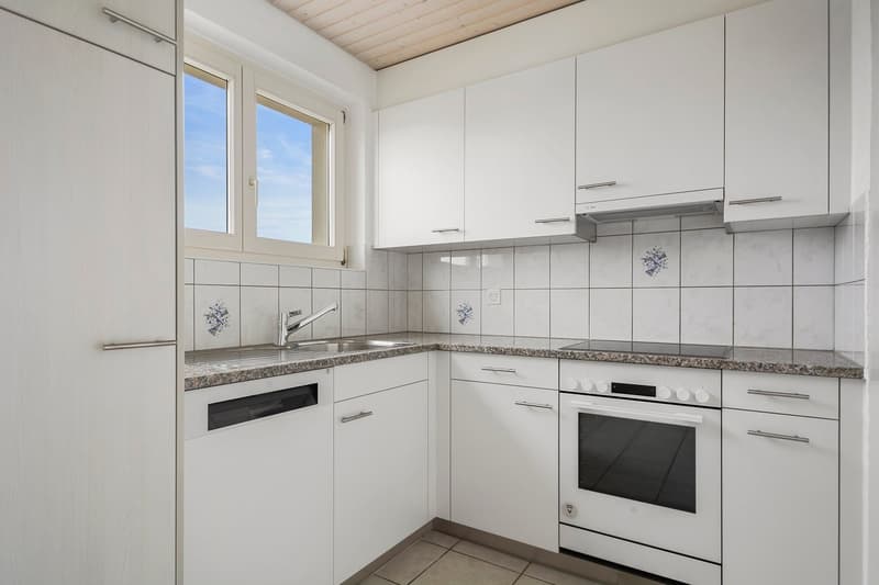Bright and modern kitchen with white cabinets, granite countertops, tiled backsplash, and stainless steel appliances including a dishwasher. Large windows provide natural light.