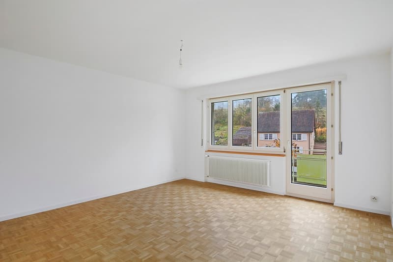 Spacious room with large windows overlooking a garden, hardwood parquet flooring, and a sliding glass door leading to a balcony or terrace.