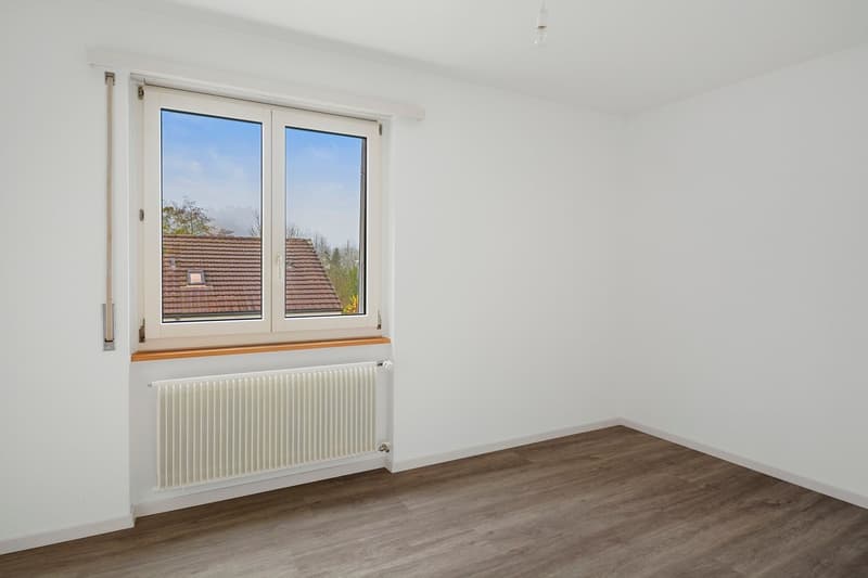 Empty room with white walls, hardwood flooring, and a large window overlooking a tiled roof and outdoor scenery.