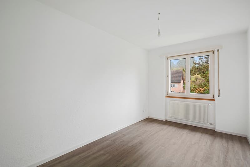 An empty room with white walls, wooden floors, a white radiator below a window, and a window with a view of trees outside.