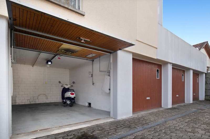 Open garage with scooter, white walls, wooden ceiling, brick floors
