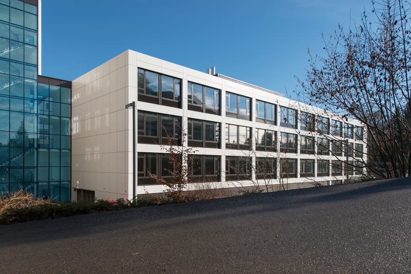 A modern, multi-story office building with a glass facade and a white exterior. The building has multiple floors and large windows. There is a paved driveway or parking area in front of the building.