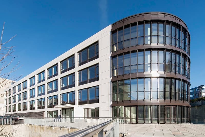 Modern multi-story office building with a curved glass facade, surrounded by a paved outdoor area with railings