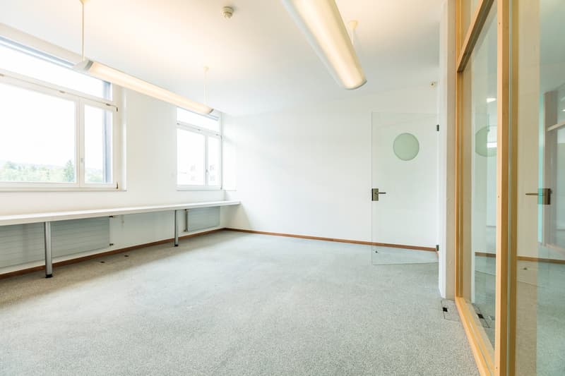 The image shows a spacious, well-lit office space with white walls, a large window, and a carpeted floor. There is a desk or workstation along the wall, and a door leading to another room. The overall atmosphere appears clean and minimalist.