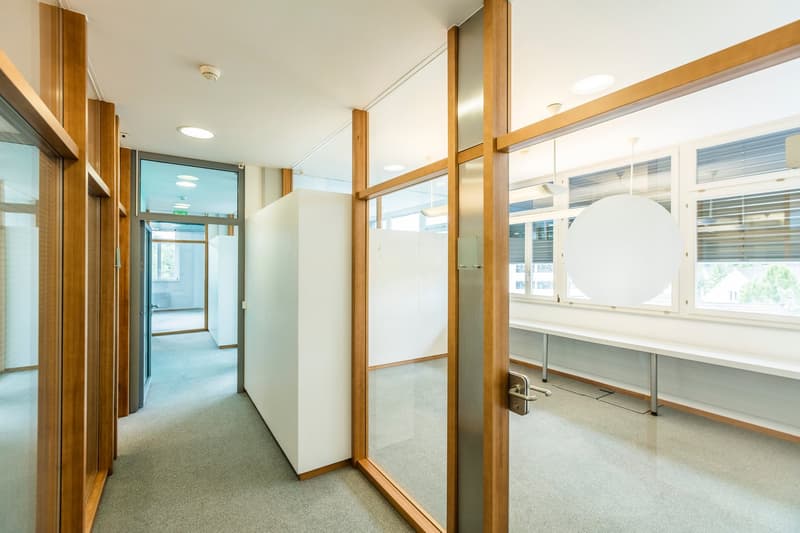 The image shows a hallway or corridor in an office or commercial building. The walls are white with wooden frames, and there are large windows allowing natural light to enter the space. The floor is covered with a light-colored carpet. The hallway appears