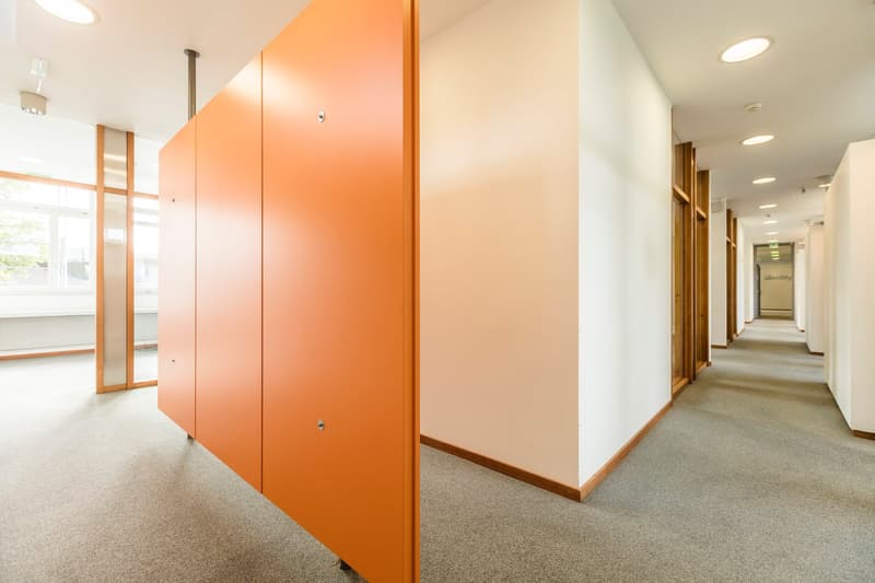 The image shows a hallway or corridor in an office or commercial building. The walls are painted in a light, neutral color, and the floor is covered with a beige carpet. There are several orange-colored doors or panels lining one side of the hallway, whic