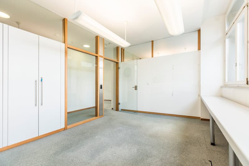 The image shows a spacious office space with white walls, a gray carpeted floor, and large glass windows and doors. The room has a minimalist design with wooden accents on the frames of the glass partitions. There is a small window ledge or counter along