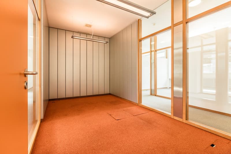 This image shows a room with an orange-colored wall, wooden paneled walls, and a large window. The room has a cork flooring and appears to be a storage or utility room with a clothing rack installed on the wall.