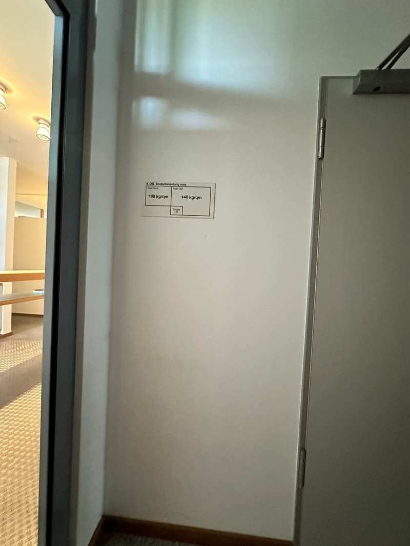 The image shows a room with a white wall and a refrigerator or appliance on the right side. There is a label on the wall indicating a maximum load capacity of 180 kg/qm or 140 kg/qm.