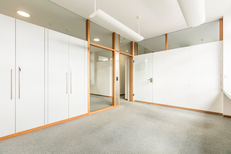 The image shows a spacious office space with white walls, wooden accents, and a carpeted floor. The room features built-in storage cabinets along one wall and large glass windows that provide natural light. The overall design appears to be modern and mini