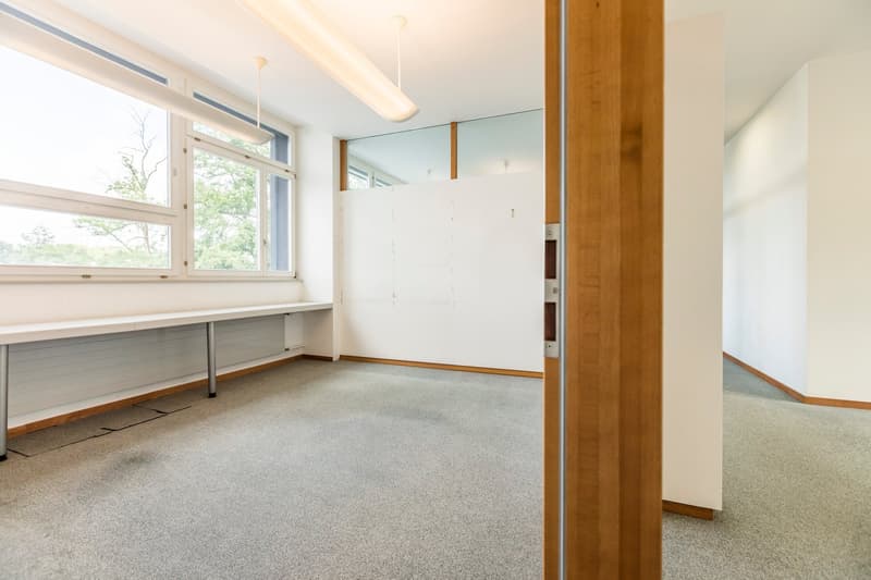 This image shows a spacious room with large windows, providing ample natural light. The room has a carpeted floor and white walls, with wooden accents and built-in shelving units along the walls. The room appears to be a study or office space, with a desk