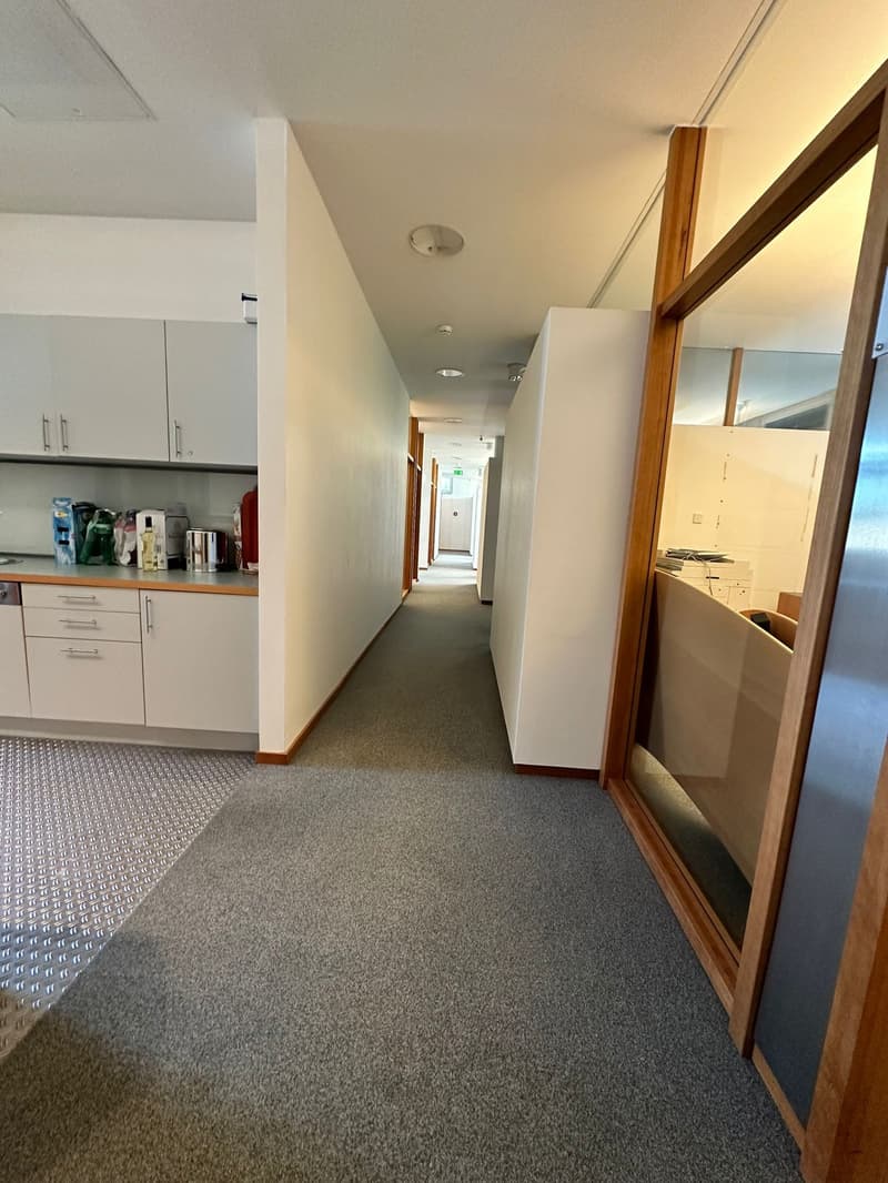 The image shows a long hallway with white walls, a gray carpeted floor, and wooden trim around the doors and windows. There is a kitchen area visible at the end of the hallway, with cabinets and appliances. The hallway appears to be in a commercial or off