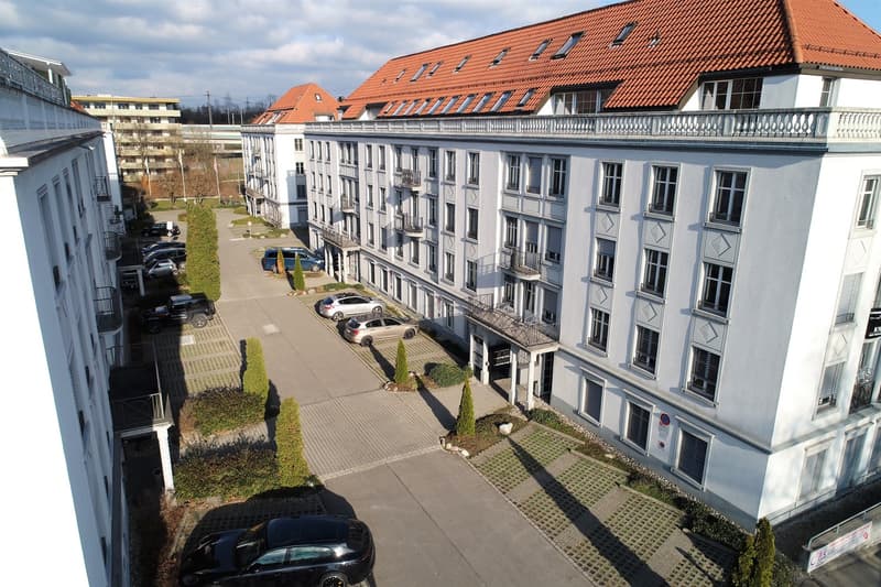 A large white building with a red roof, many windows, several floors, and a parking lot in front.