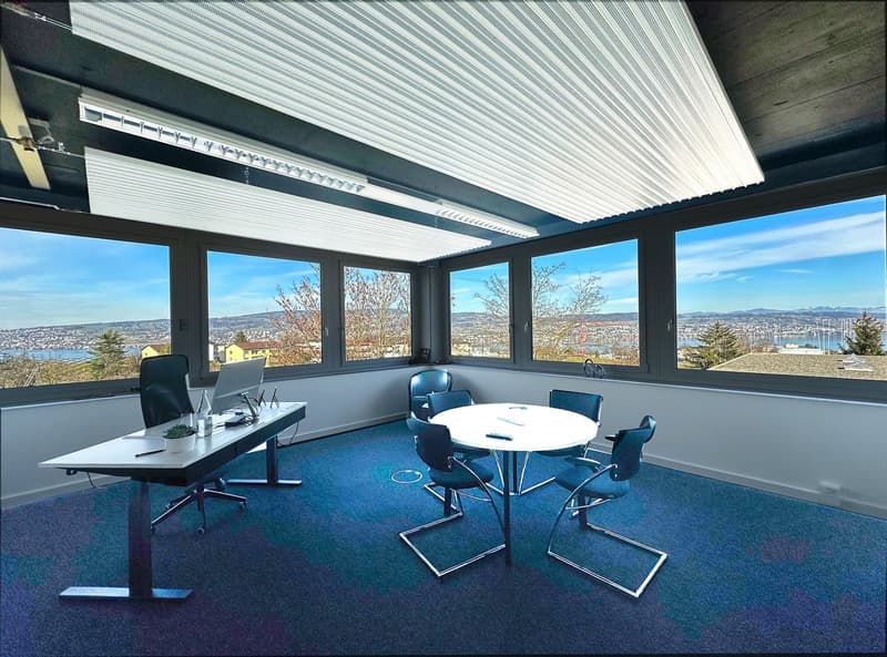 Modern office space with large windows offering a scenic view, a round meeting table with blue chairs, and a desk with a laptop and other office equipment.