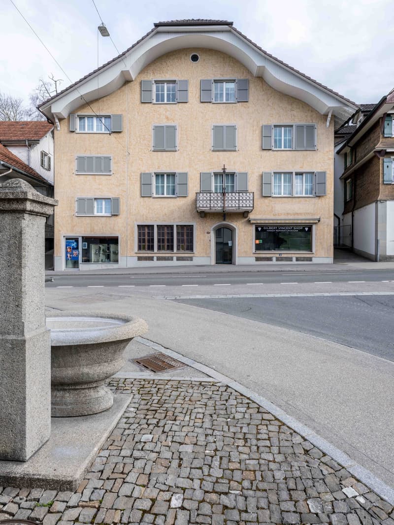 Building with tiled facade, multiple windows, balcony, concrete fountain, cobblestone sidewalk