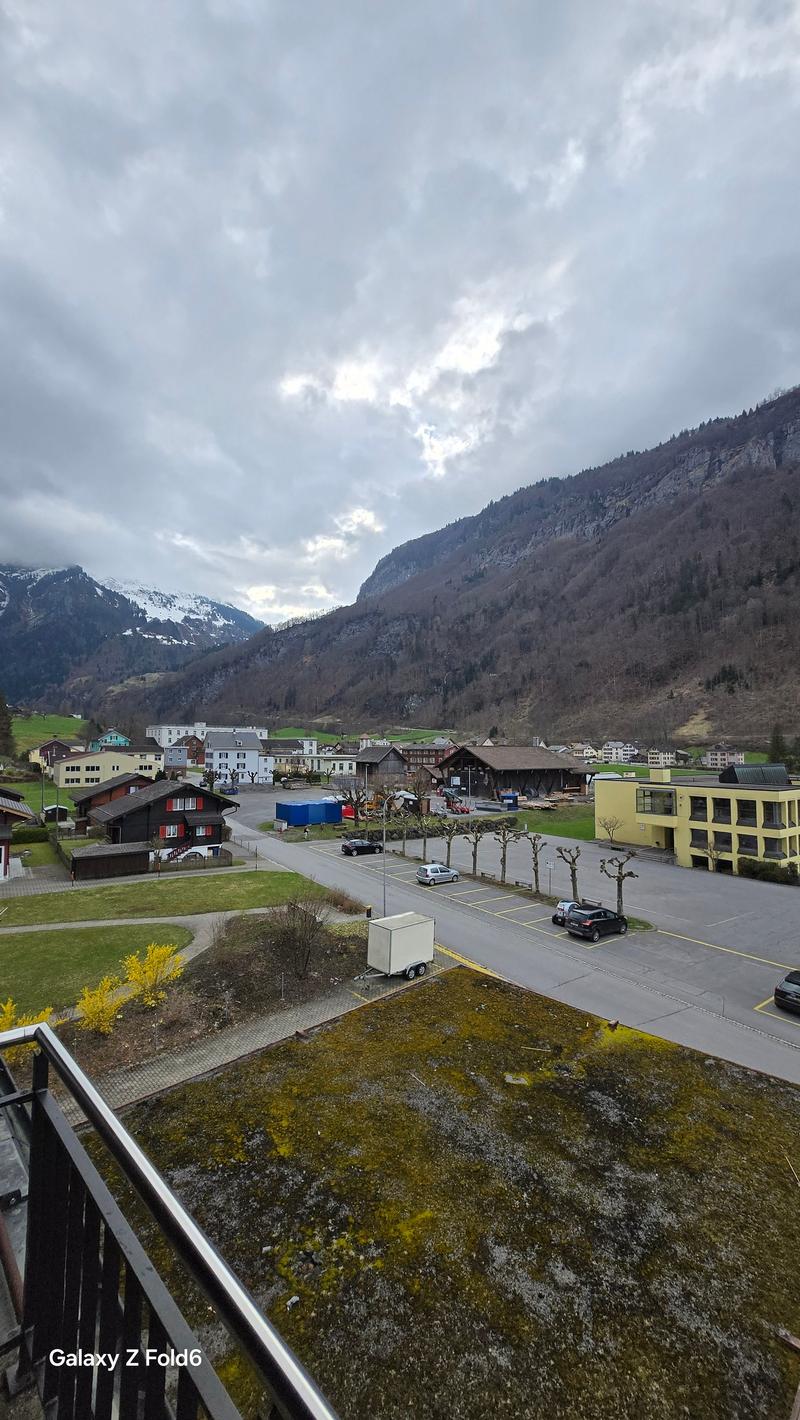 Aerial view of a parking lot with several cars parked, a grassy area, trees, and a building in the distance. Mountains and a cloudy sky in the background.