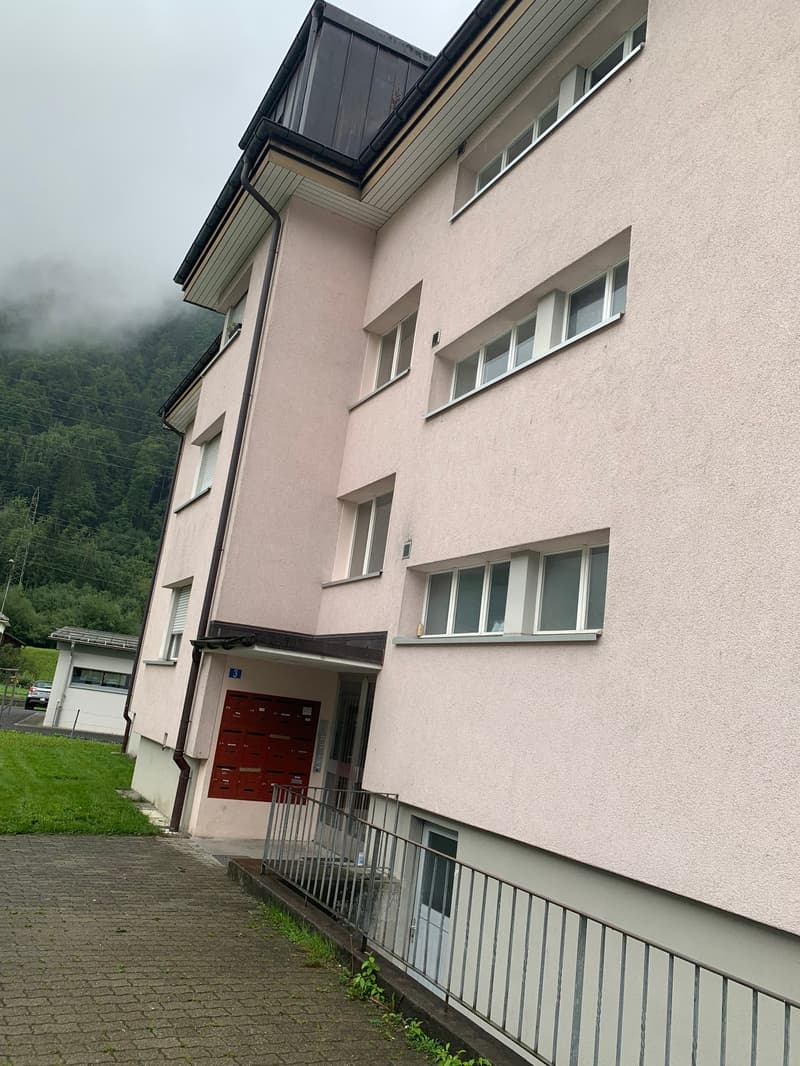 4 story building, pink exterior, multiple windows, metal railing, red mailbox, paved walkway, small grassy area, mountain in the background