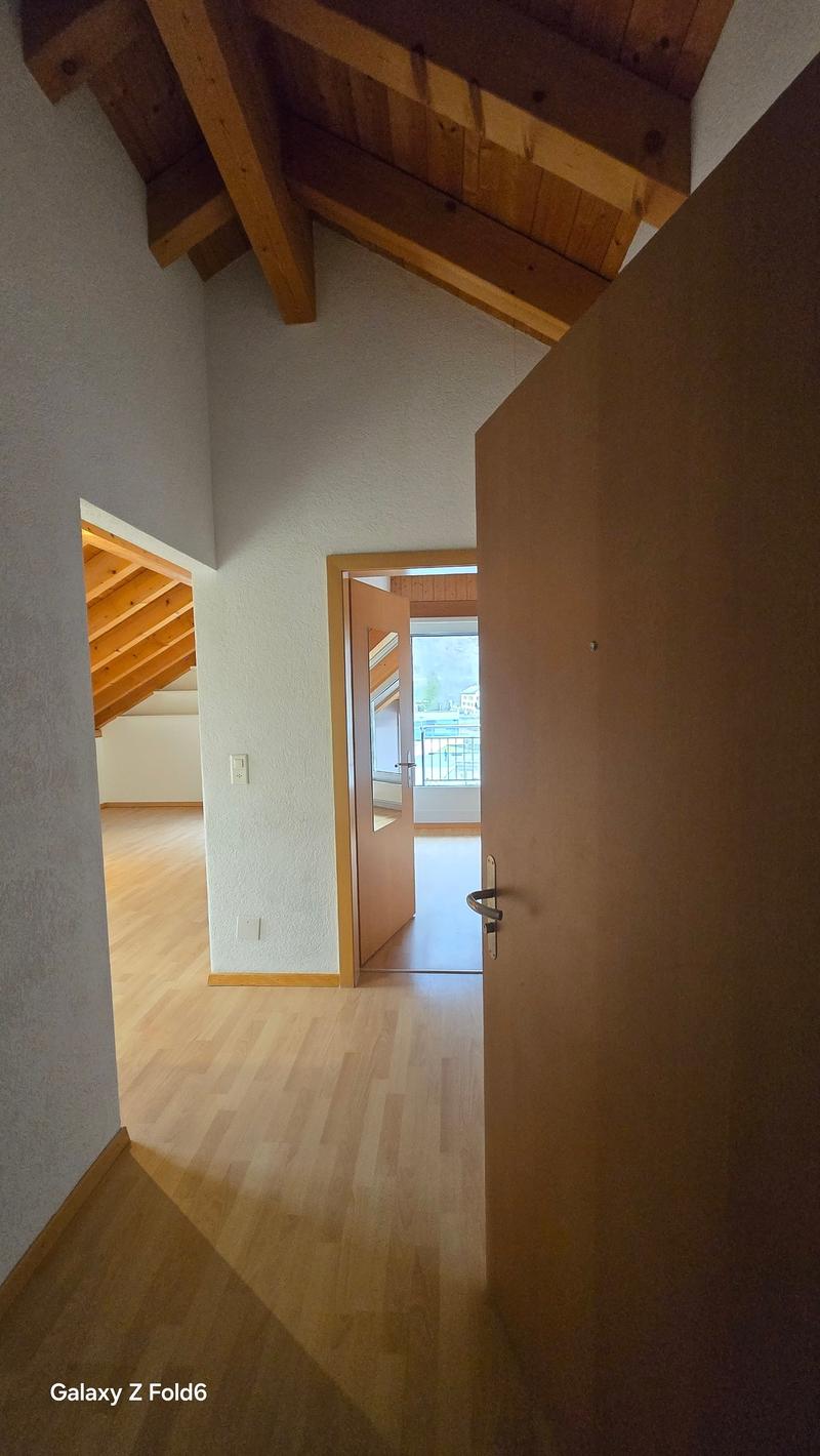 The image shows the interior of a room with a high, slanted wooden ceiling and wooden beams. The floor is made of light-colored hardwood. There is a doorway visible, leading to another room or outdoor space. The walls are painted a neutral color.