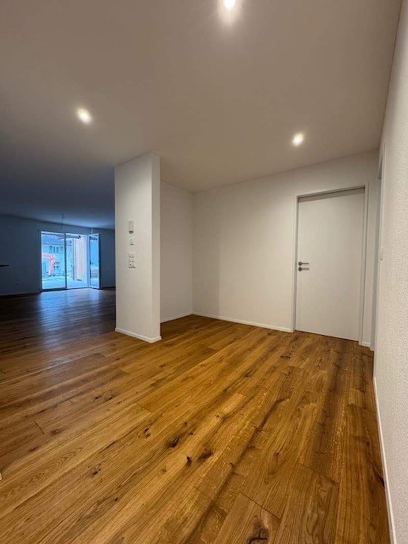 An empty room with hardwood flooring, white walls, and white doors. There are windows and sliding glass doors that provide a view outside.