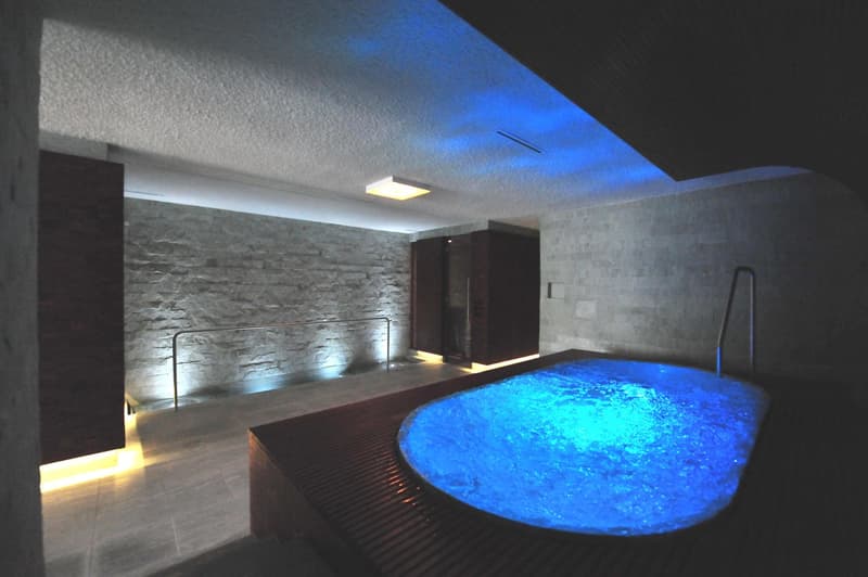 Indoor pool with blue lights, adjacent wooden bench, light on ceiling, wall has yellow lighting