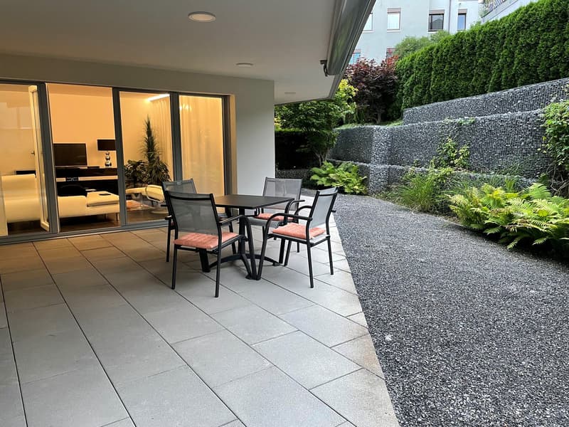 The image shows a patio or outdoor living area adjacent to a modern apartment or home. The patio has a tiled floor and is furnished with a dining table and chairs. The outdoor area is surrounded by lush greenery, including shrubs and trees, creating a pri
