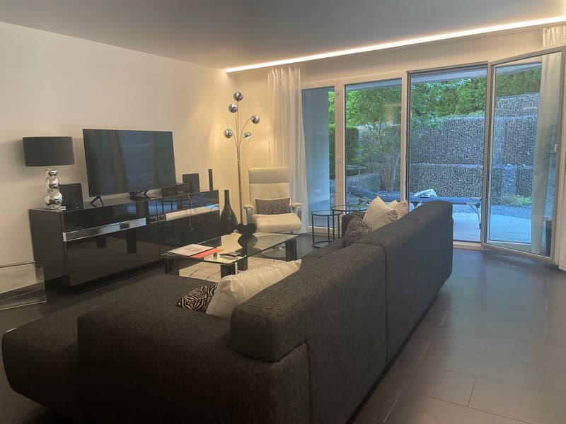 Spacious living room with large windows overlooking a balcony or terrace, dark gray sofa, black entertainment unit with decor, and modern lighting fixtures.