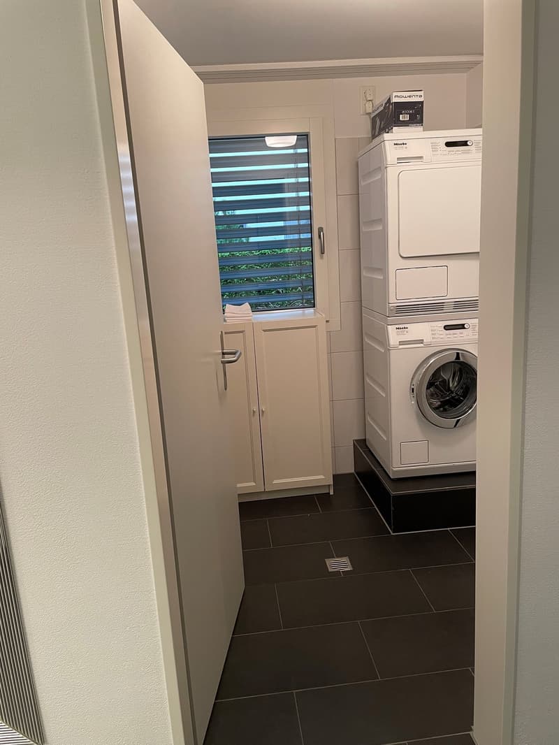 The image shows a laundry room with a washing machine and a dryer stacked on top of each other. There is also a dishwasher visible in the room. The room has a tiled floor and a window with blinds that provides natural light.